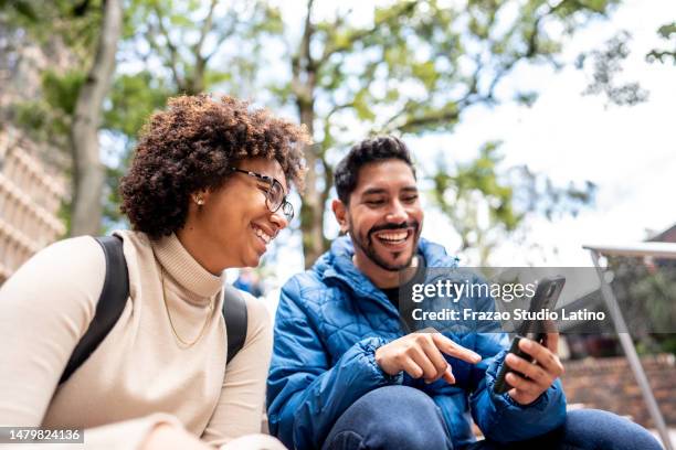 young man showing something on mobile phone to his friend on stairs outdoors - two friends laughing stock pictures, royalty-free photos & images