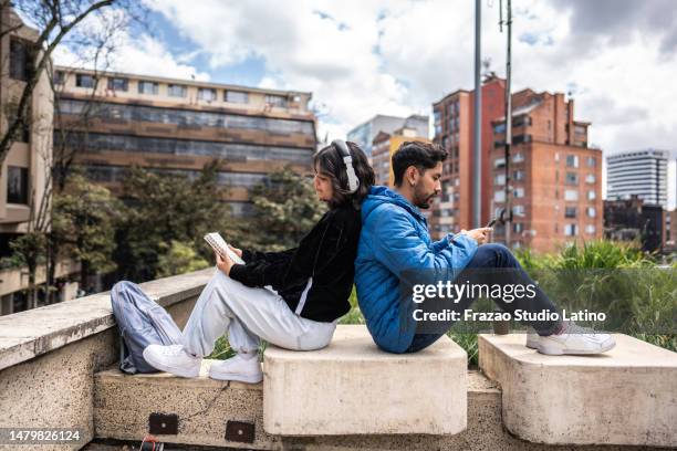 young man using mobile phone while his friend read a book outdoors - back to back leaning stock pictures, royalty-free photos & images