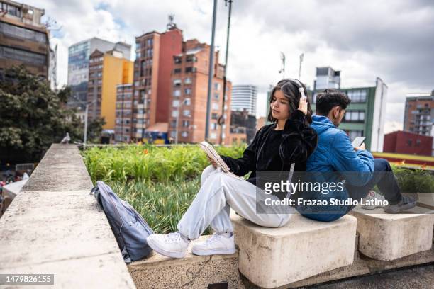 young woman listening music while read a notepad leaning against her friend outdoors - back to back leaning stock pictures, royalty-free photos & images