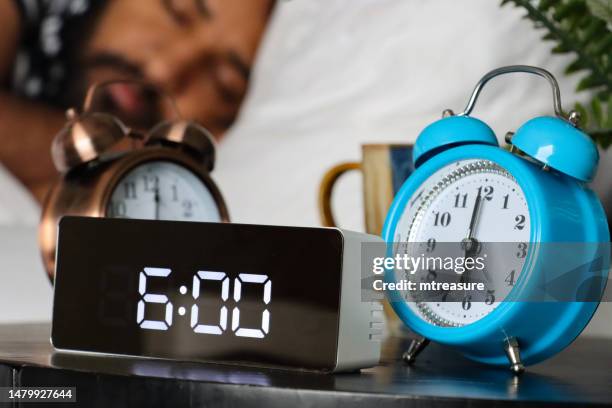nahaufnahme von drei weckern auf dem nachttisch, die auf 06:00 uhr eingestellt sind, schlafender mann am frühen morgen für die arbeit, weiße bettdecke und bettwäsche, digitale und analoge zeit, blaue und metallische uhren - uhrzeiger stock-fotos und bilder