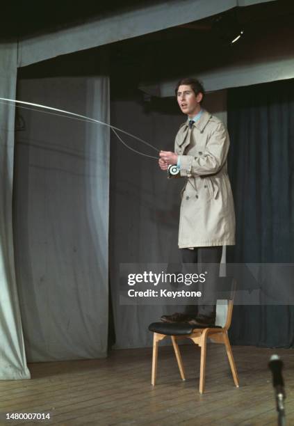 British Royal Charles, Prince of Wales, wearing a raincoat during a sketch for the Trinity College revue sketch, in which he stands on a chair...