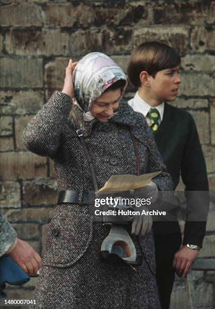 British Royals Elizabeth II wearing a headscarf and a grey tweed coat, scratches her head, with a Leica M3 camera hanging around her neck, with her...