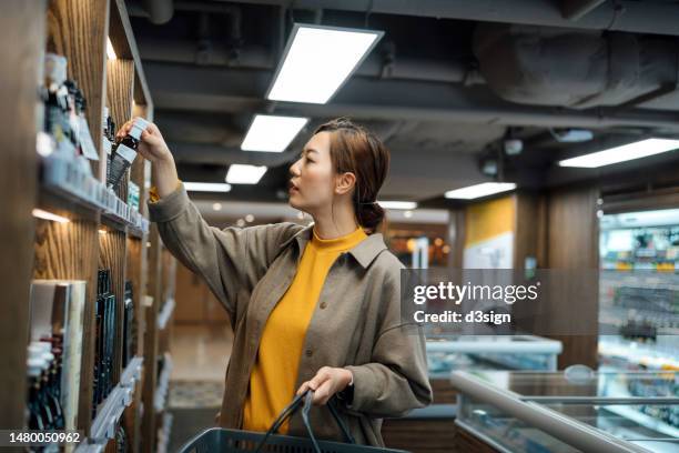 young asian woman carrying a shopping basket, grocery shopping in supermarket, choosing a bottle of cooking sauce from the shelf. food shopping. making healthier food choices - shopping basket bildbanksfoton och bilder