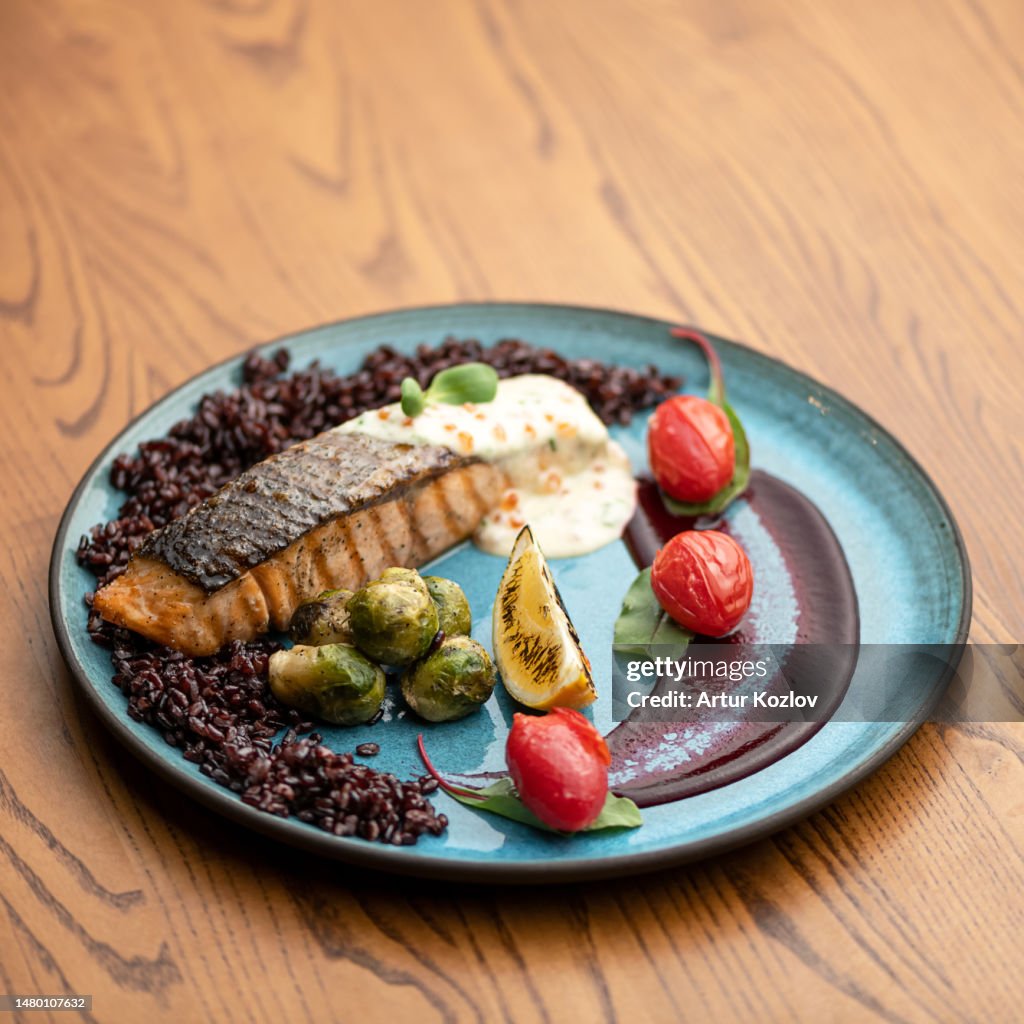 Salmon fillet with garnish and sauce on blue plate. Grilled fish fillet with brown rice, baked Brussels sprouts, cherry tomatoes and white sauce. Healthy food. Side view. Wooden background. Copy space