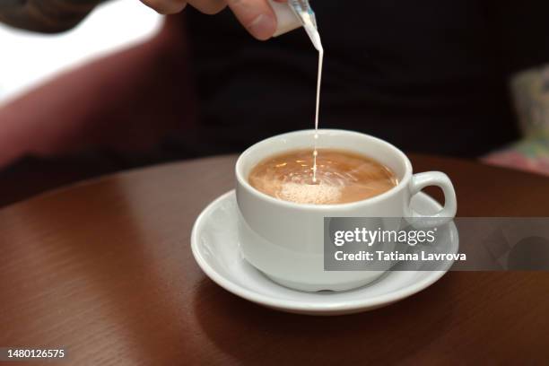 in the cozy teahouse. male hand pouring milk into black tea in the white ceramic cup on saucer. closeup of natural hot beverage. front view, high angle - english tea room stockfoto's en -beelden