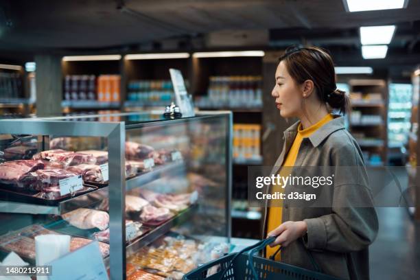 young asian woman choosing some fresh meat at meat section in the supermarket. food and grocery shopping. eating well. choosing quality and healthy source of food in environmentally conscious way. healthy eating lifestyle - mercado espaço de venda no varejo - fotografias e filmes do acervo