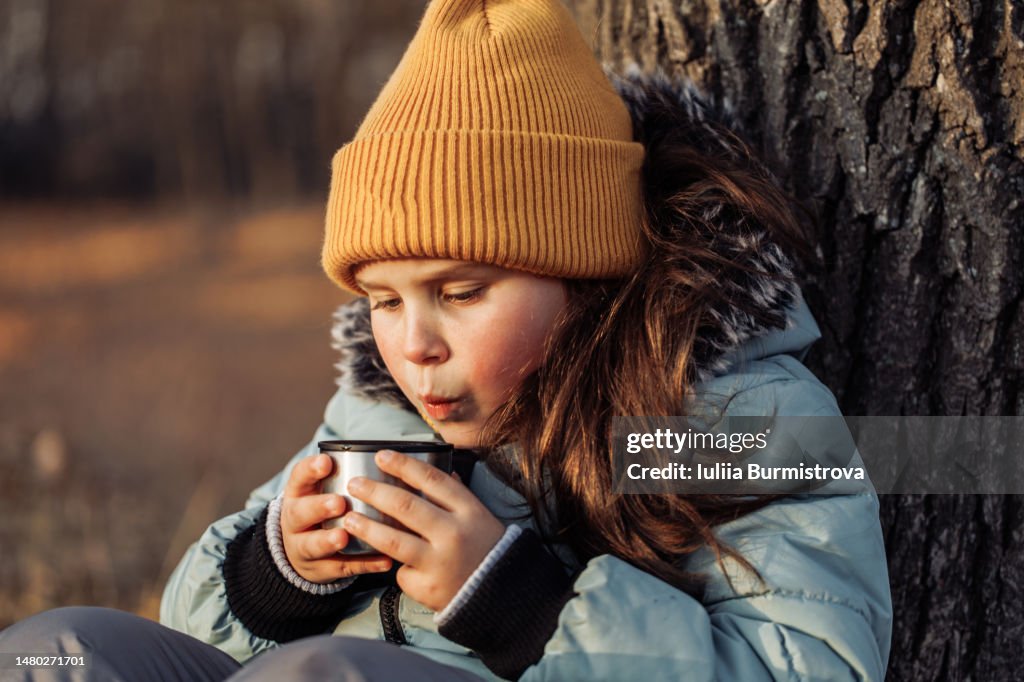Portrait of school aged girl holding cup of hot tea from thermos and blowing with mouth. Closeup, selective focus.