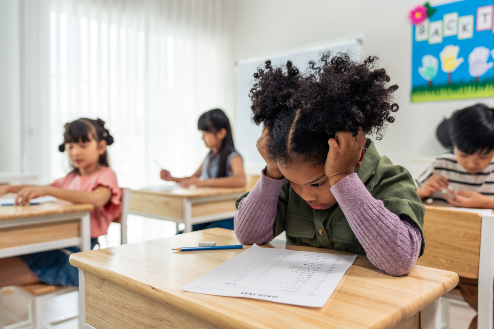 African American black girl student doing an exam at elementary school. Adorable young girl children sitting indoors on table, feeling upset and depressed while learning with teacher at kindergarten. African American black girl student doing an exam at elementary school. Adorable young girl children sitting indoors on table, feeling upset and depressed while learning with teacher at kindergarten.