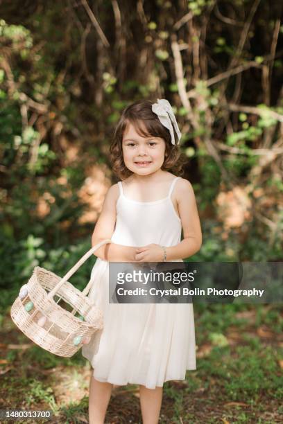 a cute cuban-american 4-year-old girl with brown curly hair & brown eyes, dressed in a cream-colored tutu dress, soft yellow floral bow, & holding a vintage egg basket, enjoying an easter egg hunt on a dreamy spring morning to celebrate the easter holiday - child picking up toys stock pictures, royalty-free photos & images