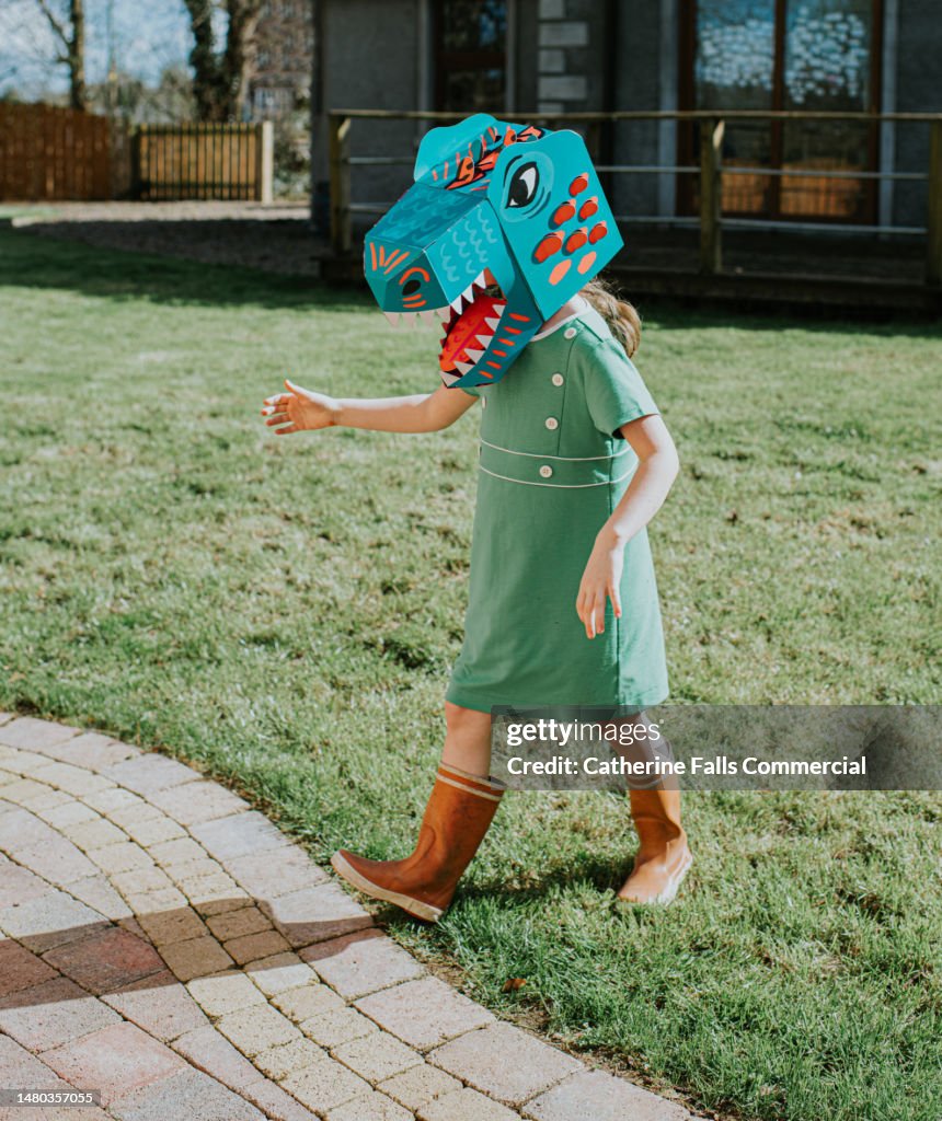 A girl in a domestic garden wears a brightly coloured cardboard dinosaur mask