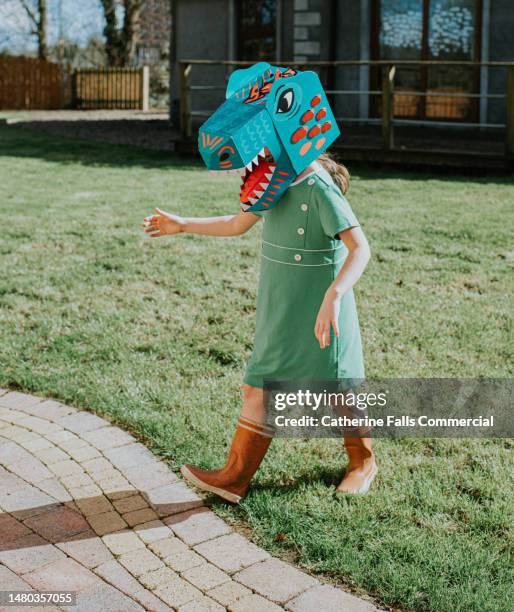a girl in a domestic garden wears a brightly coloured cardboard dinosaur mask - disguise face stock pictures, royalty-free photos & images