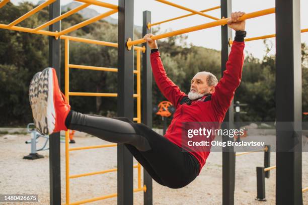 senior athletic man doing abs workout on outdoor sports ground. lifting legs on horizontal bars - senior sport stock-fotos und bilder