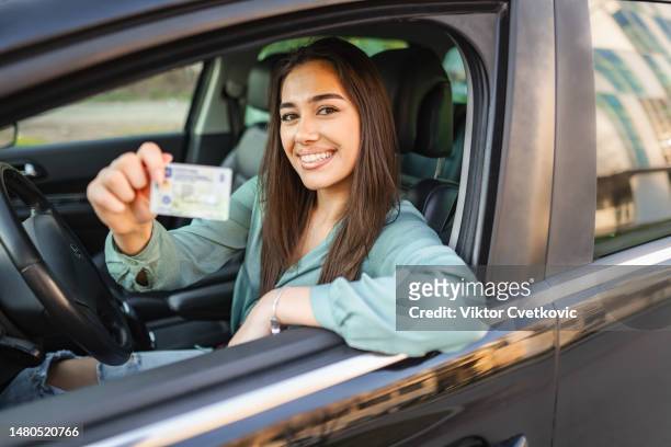 portrait of a smiling young woman sitting in the drivers seat holding drivers license - rijbewijs stockfoto's en -beelden