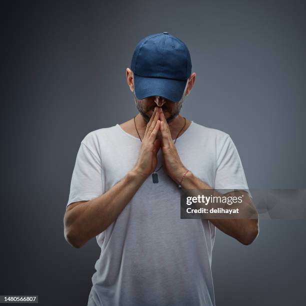 handsome man wearing white t-shirt and baseball cap, hands folded in prayer against gray background - baseball tee stock pictures, royalty-free photos & images