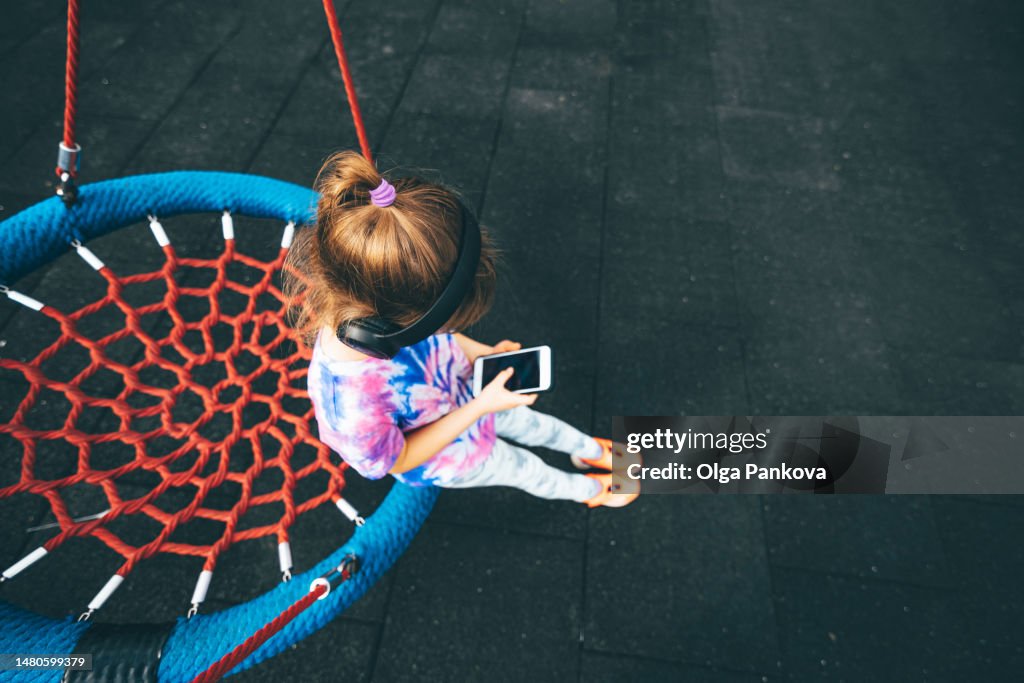 Unrecognizable young girl uses a phone and headphones sitting on a swing on a swing