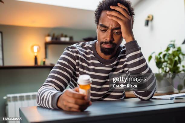 portrait of a young man sitting at the table and holding a pill bottle - confused patient stock pictures, royalty-free photos & images