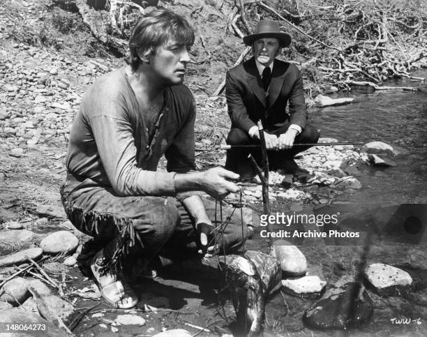 Robert Mitchum and Kurt Douglas squatting down by a river in a scene from the film 'The Way West', 1967.