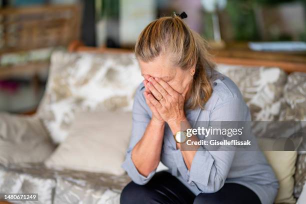 senior woman with depression sitting with her head in her hands at home - distraught elderly stock pictures, royalty-free photos & images