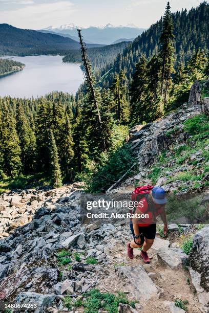 a female backpacker hikes up a steep trail with an alpine lake in the background - vancouver stock pictures, royalty-free photos & images