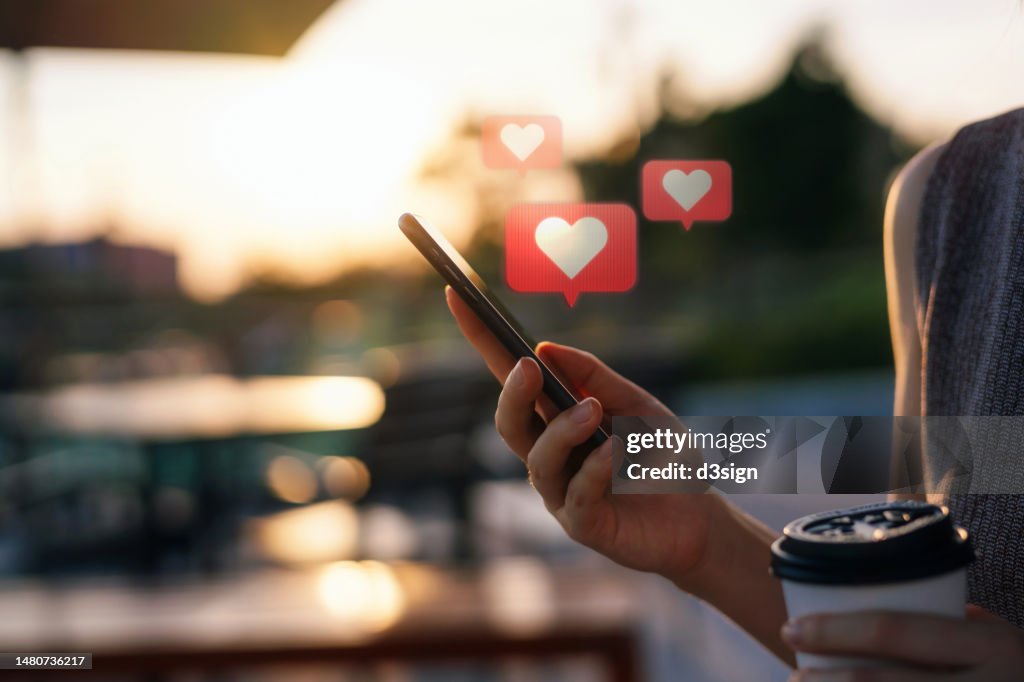 Close up of young woman checking social media network on smartphone on the go in the city. Mobile phone with heart emoji speech bubble message on the screen. People engaging in networking with technology. Social media addiction concept