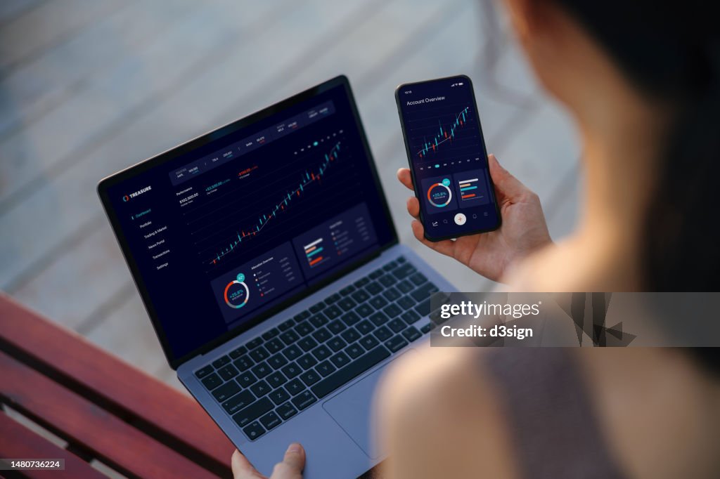 Over the shoulder view of Asian businesswoman managing finance and investment online, analyzing stock market trades and making financial plan with laptop and smartphone. Online banking. Banking, finance, investment and financial trading concept
