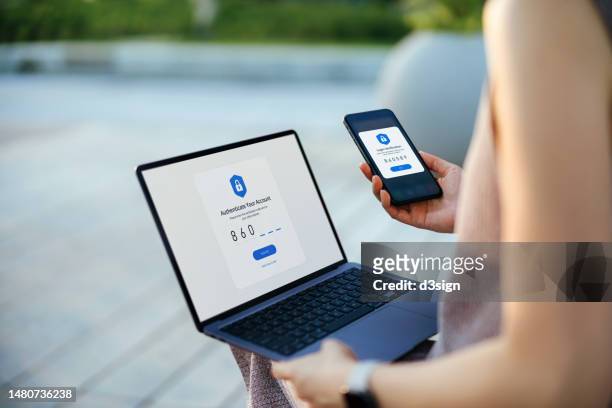 over the shoulder view of young asian woman using mobile device with two-factor authentication (2fa) security while logging in securely to her laptop outdoors in the city. privacy protection, data protection, internet and mobile security - authentication stock-fotos und bilder