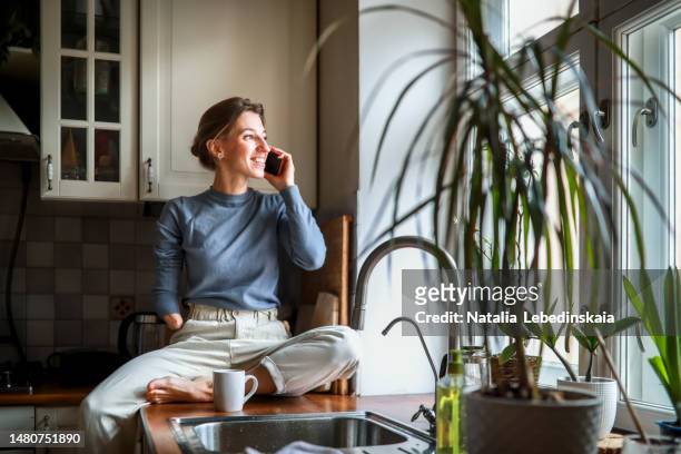 woman with disability deformed arm talking phone and looking out window on kitchen at home. relaxed happy woman in cozy apartment - disabilitycollection stock pictures, royalty-free photos & images