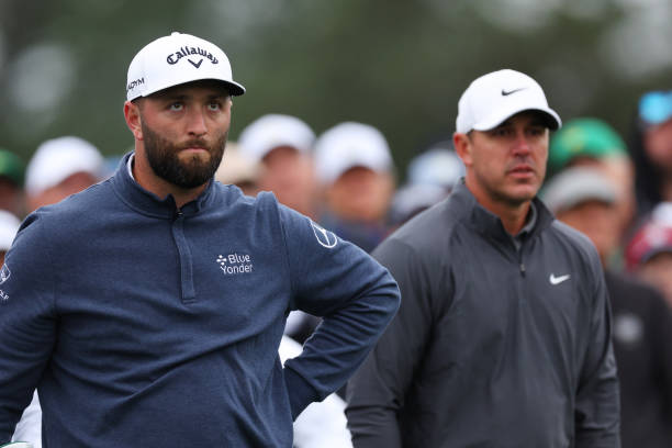 Jon Rahm of Spain and Brooks Koepka of the United States look on from the eighth tee during the continuation of the weather delayed third round of...