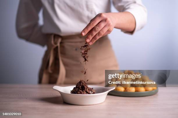 female chef sprinkling chocolate chip on a bowl of profiteroles with chocolate sauce - strooisels stockfoto's en -beelden