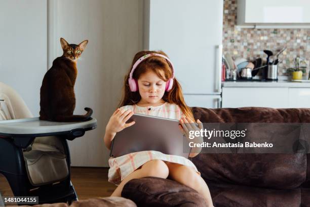 red head girl wearing head phones and using tablet. abyssinian cat sitting next to her - abyssinian cat stock-fotos und bilder