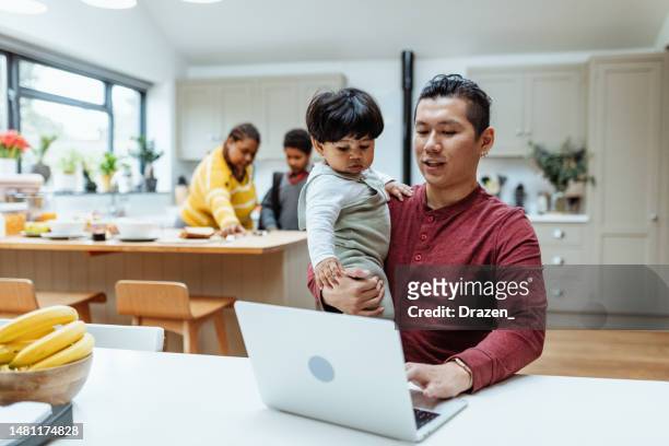 asian entrepreneur father working from home on laptop and holding baby - working from home kids stock pictures, royalty-free photos & images