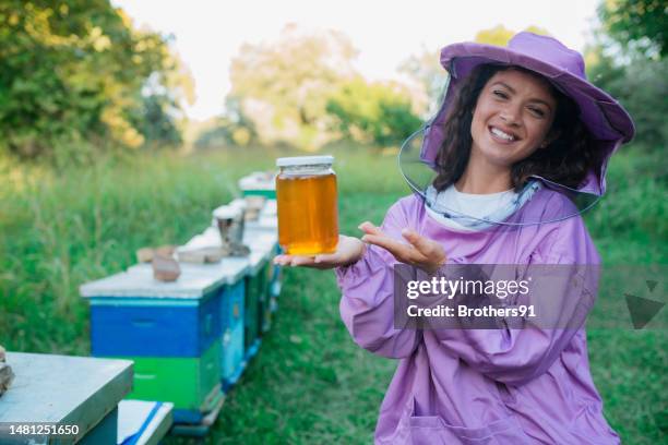 portrait of happy young female beekeeper presenting a jar of honey at apiary - honey bee stock pictures, royalty-free photos & images