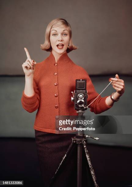 Posed studio portrait of a female fashion model wearing an orange high necked knitted cardigan, she operates a Rolleiflex twin-lens reflex camera...