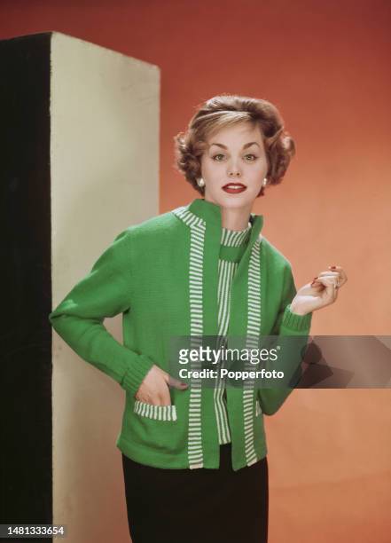 Posed studio portrait of a female fashion model wearing a green knitted twinset comprising a green and white striped tank top, and a plain green...
