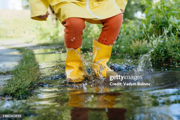 little girl in yellow boots and a raincoat jumps in a large puddle against the background of plants - puddle bildbanksfoton och bilder
