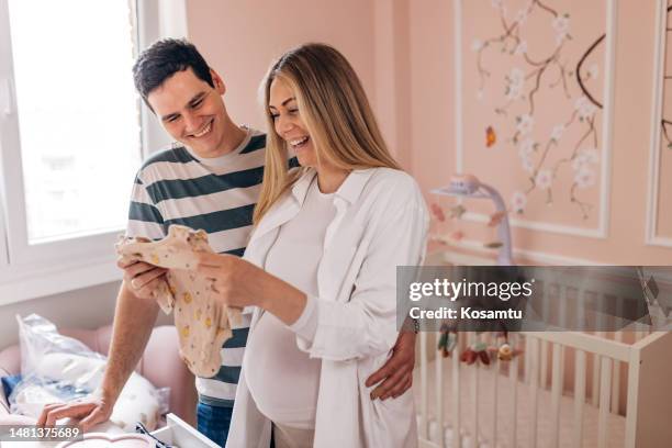 una mujer embarazada sonriente sosteniendo un armario para bebés en sus manos mientras está de pie en los brazos de su esposo en la habitación del bebé - baby shower fotografías e imágenes de stock