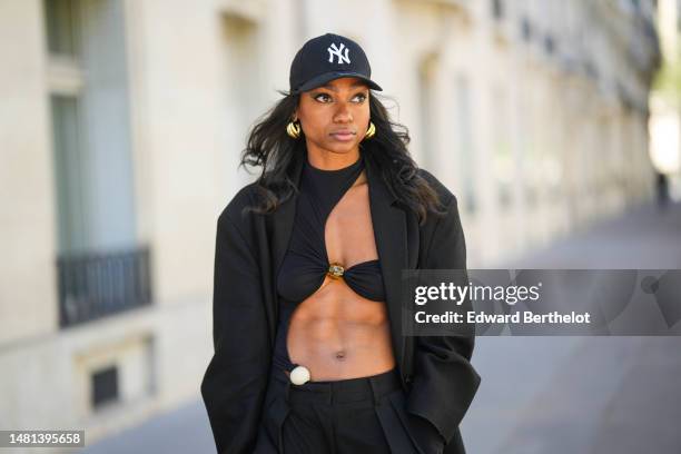 Emilie Joseph wears black with white embroidered NY logo pattern cap from New Era, black sunglasses, gold large earrings, a black cut-out asymmetric...