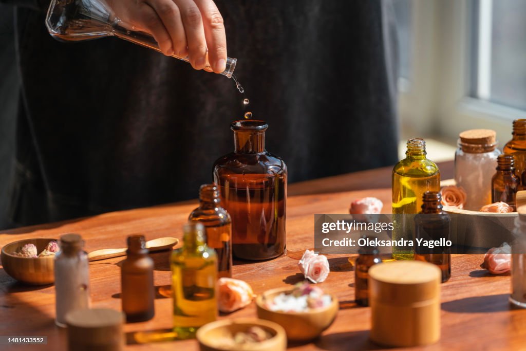Woman preparing aromatic liquid for diffuser on wooden table. Many bottles and jars with different types of oils, salt and essences. Alternative medicine concept.