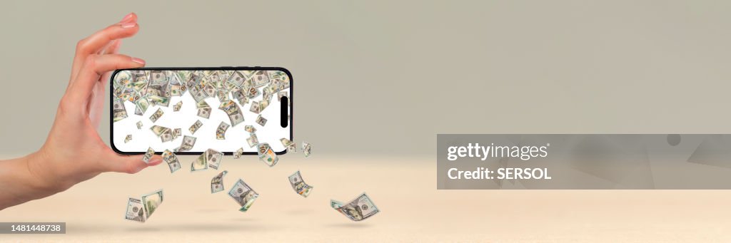 Money is falling from a mobile phone, a hand is holding a phone, US dollars are pouring out of the phone. Financial investment concept from mobile app.