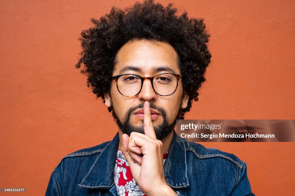 Latino non-binary person with glasses and afro hairstyle with a finger on his lips, asking for quiet, silence