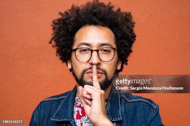 latino non-binary person with glasses and afro hairstyle with a finger on his lips, asking for quiet, silence - mystery fotografías e imágenes de stock