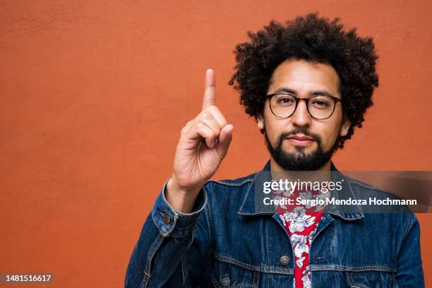 latino non-binary person with glasses and afro hairstyle giving the number one sign - number one finger sign stock pictures, royalty-free photos & images