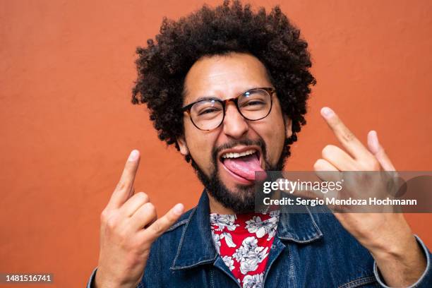 latino non-binary person with glasses and afro hairstyle doing the heavy metal horns hand sign, sticking out their tongue - heavy metal horns stock pictures, royalty-free photos & images