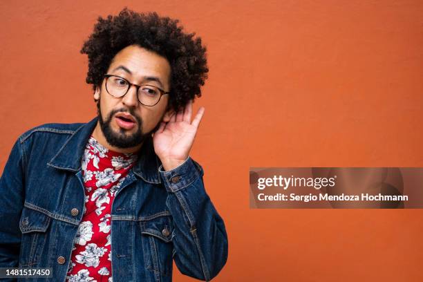 latino non-binary person with glasses and afro hairstyle putting his hand to his ear and listening intently - listening intently stock pictures, royalty-free photos & images