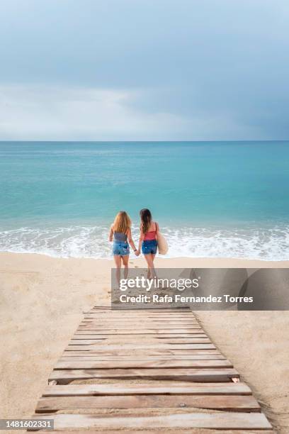 two female friends in jeans shorts walking hand in hand on the beach - woman swimsuit happy normal stock pictures, royalty-free photos & images