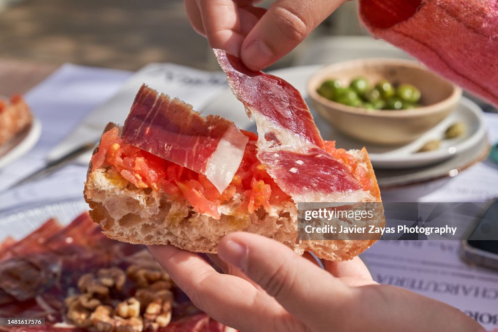 Hand putting serrano ham on top of a slice of toasted bread with crushed tomato on top