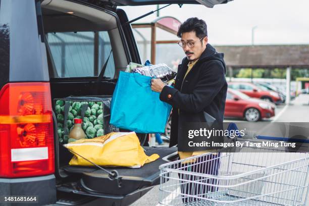 young asian man putting away bags of groceries in his car trunk at supermarket car park - supermarket uk stock pictures, royalty-free photos & images