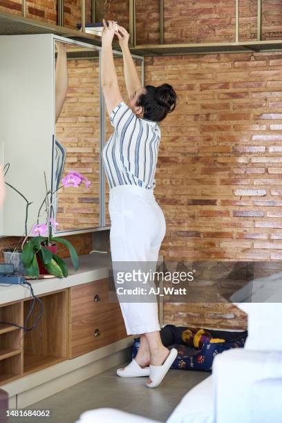 young woman reaching for a book on a shelf in her living room - reaching higher stock pictures, royalty-free photos & images