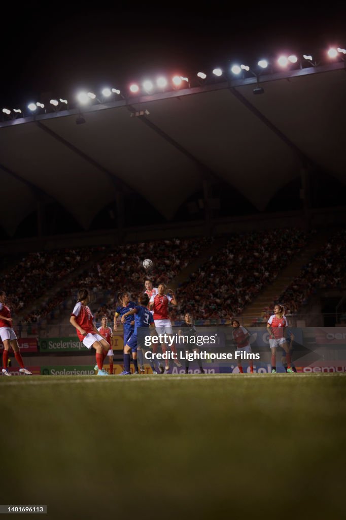 Two opposing soccer teams go after a soccer ball in mid-air in front of a crowded stadium.