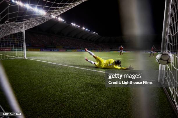 action rear view of a female soccer goalie diving and narrowly missing the ball before it goes in the goal - football goal stock pictures, royalty-free photos & images